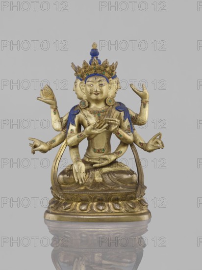 Ushnishavijaya, 19th century, South China, / Tibet, sculpture, height 17.0 cm, width 13.5 cm. Depicts Ushnishavijaya on a lotus throne, statuette with mudras; Buddhist iconography including ushnisha; materials and, An identical figure is in another museum., A gilded multi-armed seated figure on a decorative base with a reflective surface below, displaying several hands in different gestures and wearing a tall ornate crown with blue and green accents, blue shoulder adornments, circular ear ornaments, and colored inlays on the torso and arms; the figure has a calm facial expression, a serene smile, and multiple faces visible from the sides, with the overall colors including gold, blue, green, and hints of red against a neutral gray background.