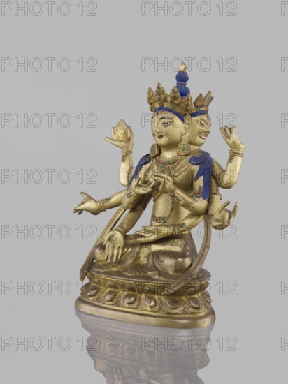 Ushnishavijaya, 19th century, South China, / Tibet, bronze sculpture, height 17.0 cm, width 13.5 cm; lotus throne, ushnisha; statuette depicting Ushnishavijaya with mudras; Buddhist iconography; inscription/attribution notes: Choynowski, Józef; Dawidow, Alexander Petrovich; An identical figure is in New York Gerald Kozicz., A gilded metal statue of a multi-armed, multi-faced seated figure on an ornate base, shown against a plain light gray background; the figure wears a crown and jewelry with blue and green enamel details, with blue accents on the headdress and shoulder coverings, and the metal surface appears gold with some darker patina; the figure’s multiple arms are arranged in various gestures, some holding small rounded objects, and the statue’s base shows carved lotus-like motifs with the whole piece reflected faintly on the glossy surface below.