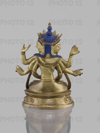 Ushnishavijaya, 19th century, South China, or Tibet, sculpture, height 17.0 cm, width 13.5 cm. Subject and iconography: Ushnishavijaya Buddhist deity with ushnisha, lotus throne, mudras; figurine/statuette in Tibetan Buddhist art; related to Buddhist art and Buddha iconography. An identical figure is in the JMMTA in New York., A small gilded statue with blue-painted hair and crown sits on a rounded base; the figure has multiple arms extended in different gestures and a three-faced head with blue head covering, all rendered in metallic gold with accents of blue and small areas of red on the faces, set against a plain light gray background.
