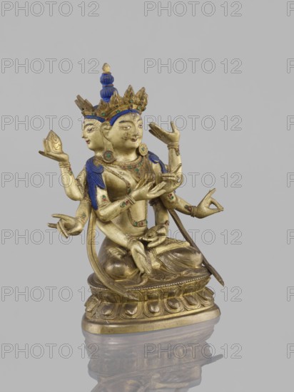 Ushnishavijaya, unknown author, 19th century, South China, or Tibet, bronze sculpture, statuette 17.0 cm high x 13.5 cm wide; lotus throne, mudras; Buddhist art iconography: Ushnishavijaya, Buddha, ushnisha. An identical figure is in New York., A gilt metal sculpture of a seated multi-armed figure with a secondary face at the back, showing a polished gold surface with blue painted hair and crown details and small green and red inlaid accents; the figure wears ornate jewelry and a tiered crown, holds various hand gestures and small objects, and sits on a lotus-style pedestal with carved petal motifs, all reflected faintly on a glossy surface beneath; visible colors include gold, blue, green, red, and a neutral gray background.