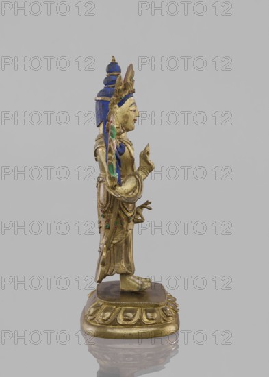 Maitreya Buddha of the Future, Tibetan Buddhist art, figurine, bodhisattva iconographic, lotus throne iconographic, Buddhist art, buddha iconographic, Buddha of the Future, 19th century, Southern China, Tibet, sculpture, height 16.8 cm, width 7.6 cm, A gilded figurative statue shown in profile standing on an ornate rounded base with a carved floral-like border, the figure wearing layered robes and a tall headdress with a tiered backpiece, painted details in blue, green, and red on the headdress and garments, the face and hands painted with flesh tones and red on the lips, the figure holding a small object near chest height with one hand while the other hand is extended forward, the base and statue surfaces showing metallic gold tones and subtle areas of wear, all set against a plain light gray background.