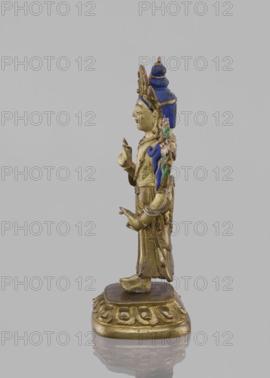 Maitreya Buddha of the Future, Tibetan Buddhist art figurine; bodhisattva on lotus throne; 19th century; Southern China, Tibet ; sculpture, height 16.8 cm, width 7.6 cm., A gilded statue of a standing figure is shown in profile against a plain gray background, with a blue headdress and blue decorative elements on the hair and garment, green and pink floral or leaf motifs on the shoulder area, and subtle red accents on the face and jewelry; the figure wears ornate jewelry and layered clothing with engraved details, holds its hands in distinct gestures, stands barefoot on a decorative gold pedestal with a rounded base and petal-like motifs, and the whole object reflects soft highlights from the lighting.