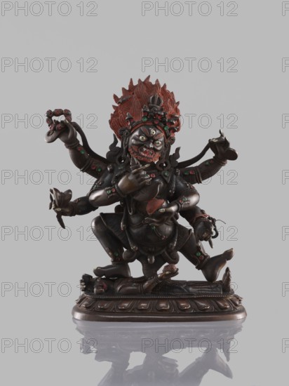 Six-armed Mahakala, dharmapala; Tibetan Buddhist sculpture, 19th century, carved figure on a lotus throne; height 16.4 cm; maker, inscriptions/creator, Tibet., A dark bronze multi-armed deity figure sits on a carved base with a reclining figure beneath, the deity shown with a fierce facial expression and bared teeth, bulging eyes, and a highly detailed headdress with flame-like ornamentation painted red; the statue has inlaid green and red accents on jewelry, limbs, and garments, and some areas of reddish paint on the face and hands; the base and figure are predominantly dark brown to black, with metallic sheen, and the background is a plain light gray.