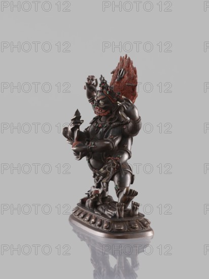 Six-armed Mahakala, dharmapala iconography; lotus throne; Buddhist/Tibetan art; 19th century . Sculpture, height 16.4 cm; Tibet., A dark bronze-colored statuette of a multi-armed figure standing on an ornate oval base is shown against a plain light gray background. The figure has a prominent sculpted mane or flame-like element at the back of its head in reddish-orange, and small accents of green and red on various parts of the body and jewelry. The figure’s limbs, hands, and ornamental elements are intricately detailed, and the base has a repeating petal-like pattern and a reflective surface beneath it that produces a faint mirrored image. The overall visible colors are dark bronze, reddish-orange, green, red, and light gray.