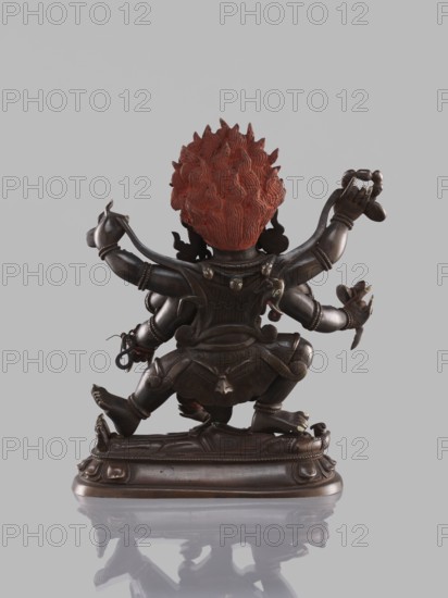 Six-armed Mahakala, dharmapala, sculpture in Tibetan Buddhist style, lotus throne iconography, possibly 19th century, height 16.4 cm, origin: Tibet., A rear view of a multi-armed bronze figure with a textured red hairpiece, seated on an ornate bronze base; the figure has several arms extended outward holding various small objects and wearing detailed armor and jewelry, with visible decorative elements on the back and limbs; the bronze shows dark brown and metallic tones contrasted with the reddish hair, and the base reflects faintly on the smooth grey surface beneath it.