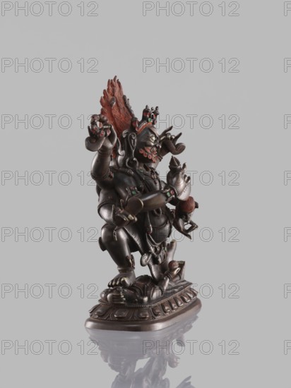 Six-armed Mahakala, dharmapala depicted seated on a lotus throne; Tibetan Buddhist sculpture, 19th century, carved/cast, height 16.4 cm., A dark metallic statue of a multi-armed figure standing on an ornate base, with a textured flame-like element rising behind its head in reddish brown; the figure has decorative accents in green and red on various parts of its body and jewelry, and small carved faces and limbs are visible among its limbs and around the base; the statue casts a subtle reflection on the pale gray surface beneath it, and the background is a uniform light gray.