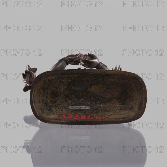 Six-armed Mahakala; creator, dharmapala iconography; lotus throne; Tibetan Buddhist sculpture, 19th century, ; material: figurine; height 16.4 cm; place of origin: Tibet., A dark brown carved object rests on a reflective surface against a light gray background, showing the underside of an elongated oval base with a weathered, textured interior that has patches of darker brown and mottled earthy tones; a pair of carved hands and floral elements in a similar dark brown wood extend from the upper edge, and there is a bright red painted mark along the lower inner rim.