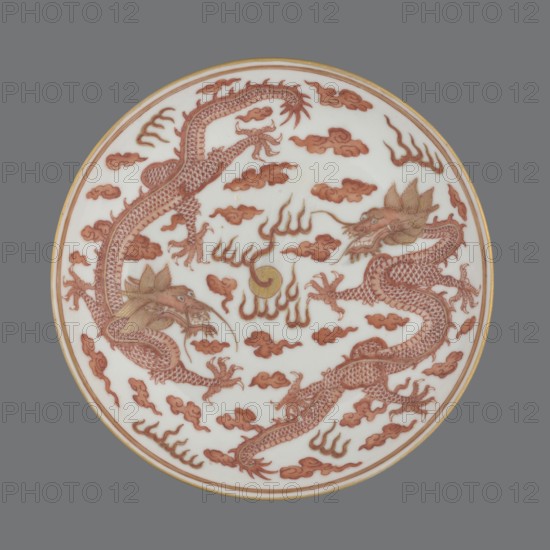 Plate., Porcelain dish decorated in the mirror and on the outer wall with a pair of dragons with a pearl among clouds on a white background; signed. Chinese; Qing dynasty, Guangxu period 1875–1908. Materials: porcelain. Height 3.2 cm; diameter 16.7 cm. Icons: dragons, pearls, clouds., A circular porcelain plate with a white background set against a gray backdrop, decorated with a pair of serpentine dragons rendered in reddish-brown with gold accents, surrounded by stylized cloud motifs in reddish-brown and flame-like shapes in reddish-brown and gold, a round golden element near the center, and a thin golden rim around the plate's edge.