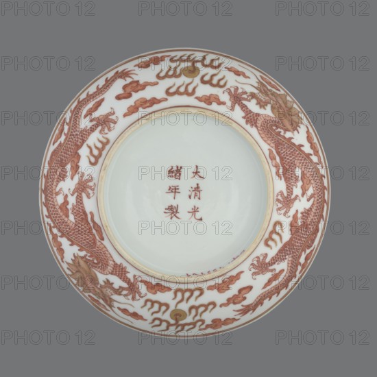 Plate., Chinese, Qing dynasty, Guangxu period 1875–1908. Porcelain dish, height 3.2 cm, diameter 16.7 cm. Decorated in the mirror and on the outer wall with a pair of dragons with a pearl among clouds on a white background. Signed., A round porcelain plate shown from above with a white central area and a glossy finish, surrounded by a decorative rim featuring a pair of stylized dragons depicted in reddish-brown with scale and claw details, set among cloud- and flame-like motifs in reddish-brown and touches of golden-yellow; the plate has a thin beige or cream inner band separating the central area from the rim, and the background behind the plate is a flat medium-gray.