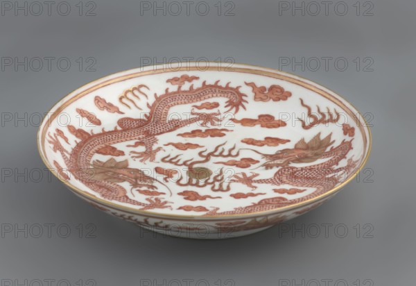 Plate, Chinese, Qing dynasty Guangxu, 1875–1908. Porcelain dish, height 3.2 cm, diameter 16.7 cm. Decorated in the mirror and on the outer wall with a pair of dragons with a pearl among clouds on a white background. Signed., A shallow round ceramic bowl sits against a gray background, showing an interior decorated with a pair of long, sinuous dragons rendered in reddish-brown tones with detailed scales, claws, and expressive open mouths, surrounded by stylized cloudlike shapes in the same reddish-brown; there is a small round element near the center with a muted greenish tone, and thin gold-colored bands rim the edge of the bowl while the bowl interior and exterior background remain white.