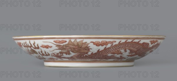 Plate. Decorated in the mirror and on the outer wall with a pair of dragons with a pearl among clouds on a white background; signed. Porcelain dish, Qing dynasty, Guangxu period 1875–1908. Dragon iconography, pearls, clouds. Height 3.2 cm; diameter 16.7 cm. Made in China., A shallow white ceramic bowl decorated with a continuous reddish-brown dragon motif and stylized cloud shapes around its exterior, with fine reddish-brown bands near the rim and base and thin gold accents along the rim; the dragon shows detailed scales, claws, a fierce open mouth, and flowing mane elements, all rendered in reddish-brown tones against the white background, set on a neutral gray surface and photographed against a muted gray backdrop.