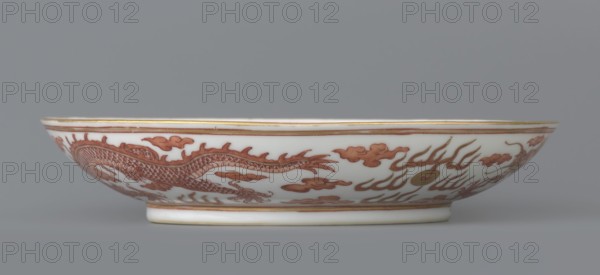 Plate, Porcelain dish, decorated in the mirror and on the outer wall with a pair of dragons with a pearl among clouds on a white background. Signed. Qing dynasty, Guangxu period 1875–1908. China. Height 3.2 cm; diameter 16.7 cm. Iconography: dragon, pearls, clouds., A shallow oval porcelain dish with a white base color and a band of reddish-brown decorative painting around its exterior showing a sinuous dragon motif and stylized cloud and flame shapes, accented with thin gold edging along the rim and base; the dish rests on a short foot and is photographed against a plain gray background.