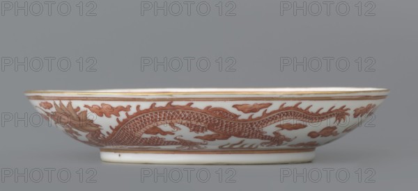 Plate, Chinese, Qing dynasty Guangxu period, 1875–1908. Porcelain dish, height 3.2 cm, diameter 16.7 cm. Decorated in the mirror and on the outer wall with a pair of dragons with a pearl among clouds on a white background. Signed., A shallow ceramic bowl photographed from the side showing its rim and exterior surface with a painted sinuous dragon motif in reddish-brown against a white background, accompanied by stylized cloud shapes in the same reddish-brown; the rim and foot of the bowl have thin gilded bands, and the overall color palette includes white, reddish-brown, gold, and subtle gray from the background.