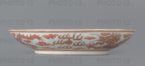 Plate. Decorated in the mirror and on the outer wall with a pair of dragons with a pearl among clouds on a white background. Signed. Porcelain dish, China; Qing dynasty, Guangxu period 1875–1908. Height 3.2 cm; diameter 16.7 cm. Iconography: dragon, pearls, clouds., A shallow white porcelain bowl viewed from the side with a low foot and a gently flaring rim, decorated with a continuous dragon motif and stylized cloud and flame shapes in shades of reddish-brown and gold, with added greenish-gold accents; the rim and foot have thin gold bands, and the background behind the bowl is a uniform gray.