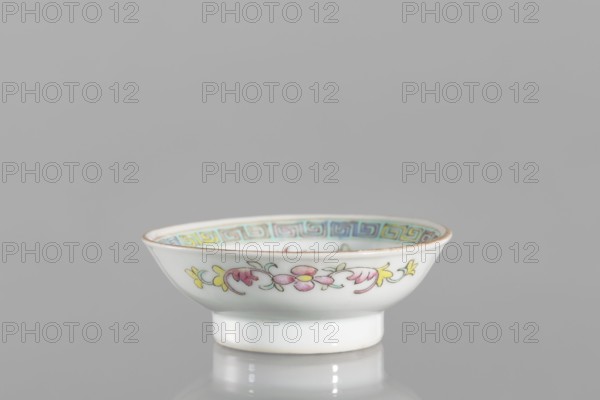 Bowl;, ; repossession Kraków; Chinese; Qing dynasty 1644–1911, Guangxu period 1875–1908; Boxer Rebellion 1899–1901 context; second half of the 19th century; colored porcelain; vessel, saucer form; floral decorations and meander ornament; height 2.7 cm, diameter 7.5 cm., A small shallow porcelain bowl sits on a reflective surface, showing a white exterior with a hand-painted floral band in pink, yellow, and pale green around its side and a narrow foot in white; the bowl rim is accented with a thin gold line and the interior rim has a decorative geometric border in shades of blue, teal, and yellow, all set against a neutral gray background.