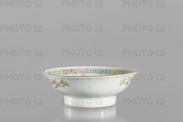 Bowl, gift, repossession Kraków; Chinese, Qing dynasty Guangxu period, 1875–1908, 2nd half of the 19th century; colored porcelain; vessel saucer, floral decorations, meander ornament; height 2.7 cm, diameter 7.5 cm; associated with Boxer Rebellion 1899–1901., A small shallow porcelain bowl sits on a reflective surface against a plain gray background, showing a white exterior with delicate painted floral motifs in yellow and pink with green accents on its sides, a faint brownish rim, and an inner border decorated with a repeating geometric band in pastel shades including blue, turquoise, yellow, and pink.