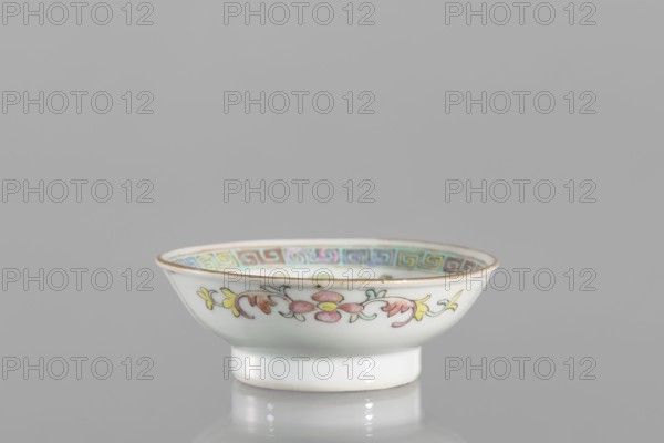 Bowl, gift; Chinese, Qing dynasty Guangxu period, 1875–1908, 2nd half of the 19th century; colored porcelain; vessel saucer with floral decorations and meander ornament; related to Boxer Rebellion 1899–1901; height 2.7 cm, diameter 7.5 cm., A small shallow ceramic bowl sits on a reflective surface against a plain grey background, showing a white exterior with a painted floral band in pink, yellow, green, and pale orange around its outer rim and a thin gold edge; the interior rim displays a decorative geometric border in muted turquoise, pink, purple, and gold, and the bowl rests on a short white foot that reflects softly beneath it.