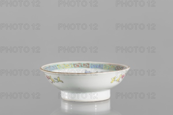 Bowl;, Colored porcelain saucer with floral decorations and meander ornament; vessel from China, Qing dynasty Guangxu period, 1875–1908, 2nd half of the 19th century. Height 2.7 cm, diameter 7.5 cm. Related historical context: Boxer Rebellion 1899–1901., A small shallow bowl with a short pedestal base sits on a reflective surface against a plain gray background, the bowl's exterior is white with painted floral motifs in yellow, pink, and green, and a narrow metallic rim. The interior rim shows a repeating geometric border in pastel shades of blue, yellow, green, and pink, and the bowl's glaze gives subtle white highlights and reflections.
