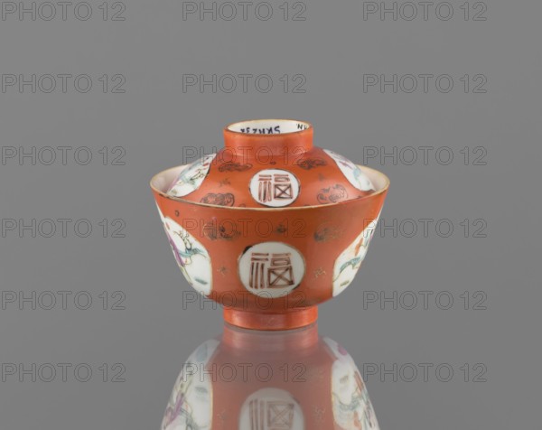 Bowl with a lid, ceramics; colored porcelain; Qing dynasty, Tongzhi period 1862–1874, China; vessel with height 8.6 cm and diameter 10.3 cm; decoration: bats and swastikas loosely placed in the background and genre scenes; inscriptions/hieroglyphs present., An orange ceramic bowl with a lid sits on a reflective surface, showing its mirrored underside; the bowl has several white circular panels around its sides and top containing painted scenes that include figures and decorative elements in colors such as green, purple, blue, and black, and the orange areas are accented with small gold decorative motifs; the background is a flat grey.