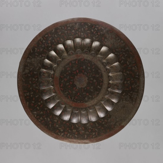 Tray, South Asian, Indian, brass tray with a separate facet; engraved and painted decoration depicting a central rosette and floral ornaments placed around it; floral and geometric ornamentation; 19th–20th century, India; vessel; height 2.0 cm, diameter 59.0 cm, A round decorative object viewed from above with a scalloped inner ring and concentric circular patterns, showing a dark, metallic surface with a mottled texture and visible patches of red and brown; the scalloped sections have a slightly reflective, silvery-grey appearance, while the surrounding areas include darker blackish-brown tones and hints of rusty red, all set against a plain light grey background.