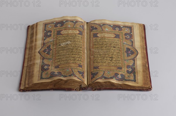 Quranic manuscript, unknown calligrapher, ; Islamic art, Ottoman dynasty 1299–1922; Arabic manuscript; written in Naskh script; illuminated; contains two pages for Surah XVII; sixteen later leaves; paper: striated, smoothed, cream, saffron-colored; 18th century, with 19th-century additions; Central Asia origin, Khiva; manuscript dimensions: height 23.0 cm, width 14.5 cm, thickness 4.2 cm., An open ornate book rests on a light gray surface, showing two facing pages with aged, yellowed paper and decorative borders. The pages feature intricate illuminated patterns in colors including gold, dark blue, green, red, and black, with elaborate floral and geometric motifs surrounding central blocks of darker script. The book cover edges visible at the sides show a worn reddish-brown leather, and the fore edges of the pages appear browned and slightly wavy from age. The spine area at the bottom center shows some wear and separation where the pages meet, and the overall scene presents a flat, over...