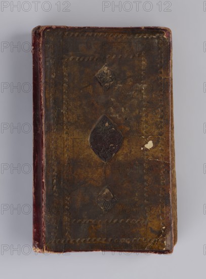 Quranic manuscript, unknown creator/calligrapher, ; Islamic art, Ottoman dynasty 1299–1922; Arabic manuscript in Naskh script; illuminated; two pages for Surah XVII; sixteen later leaves; paper striated, smoothed, cream and saffron-colored; produced 18th century with 19th-century additions; Central Asia, Khiva; manuscript; height 23.0 cm, width 14.5 cm, thickness 4.2 cm., A worn rectangular object with a dark brown surface showing a mottled texture and patches of lighter brown, featuring a central dark embossed decorative shape and two smaller embossed shapes aligned vertically, surrounded by a border of impressed curved motifs; the left edge has a vertical strip of deep red with scuffed lighter areas, and the right edge shows lighter brown wear and small areas of chipped material revealing paler tones; the background is a soft light gray.
