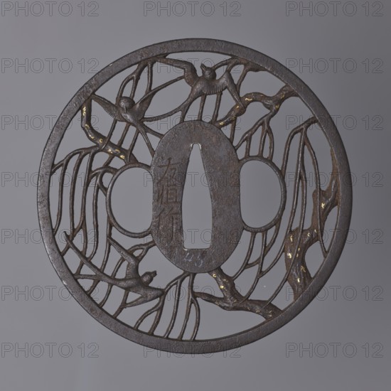 Crossguard tsuba by Nakai Tomotsune II. Early 18th century Edo period, 1603–1868. Iron round tsuba with oval seppa-dai; diameter 8.3 cm, thickness 0.4 cm. Decorated in positive openwork depicting swallows among willow branches with small gold spots nunome technique on the branches. Rectangular edge kaku mimi. Hitsu- and kogai of an identical shape. Signed. Origin: Hagi, Nagato, Japan., A round dark metal object with an openwork design sits against a neutral gray background, showing a circular outer rim and an inner raised central section with an elongated central opening flanked by rounded openings. The open spaces are filled with a network of thin branching elements that resemble twigs or vines, and a group of stylized birds are integrated into the branches, some perched and some with wings extended. The metal surface is predominantly dark brown to black with patches of lighter brown and small areas of muted gold highlights on parts of the branches. The overall appearance is flat and ...