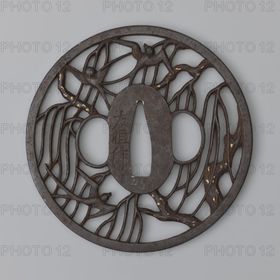 Crossguard tsuba by Nakai Tomotsune II. Early 18th century Edo period, 1603–1868. Japan — Nagato province, Hagi. Round tsuba with oval seppa-dai; hitsu- and kogai of identical shape. Decorated with positive openwork depicting swallows among willow branches. Small gold spots nunome technique on the branches. Rectangular edge kaku mimi. Signed. Diameter 8.3 cm; thickness 0.4 cm., A round dark brown metal disk with an openwork design, featuring an oval central plate with a vertical cutout and two rounded side openings; the openwork shows a group of stylized birds perched among winding branches and elongated leaves, with small areas of gold-colored spotting on parts of the branches; the metal surface has a weathered, slightly textured appearance, and the background is a pale gray.