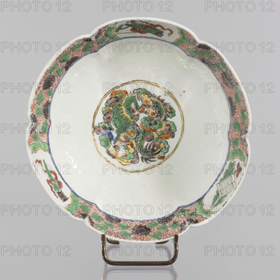 Bowl, ceramics, figural scenes, knightly scenes, Chinese art, Chinese culture, colored porcelain, backdated signatures, six-leafed, bottled belly. Porcelain body; white, transparent glaze. Decoration painted with overglaze enamel paints and iron red. 19th century, China, vessel. Height 6.5 cm, diameter 20 cm., ., A shallow scalloped ceramic dish on a metal stand with a white interior and exterior, featuring a central circular medallion containing a stylized creature and cloudlike forms in green, orange, yellow, gray, black, and a touch of blue, surrounded by a thin gold ring; the rim area has a decorative border with a repeating floral and leaf pattern in green, orange, gray, black, and pinkish-red, interspersed with several framed motifs that include similar colors, and a thin blue inner band just inside the border.