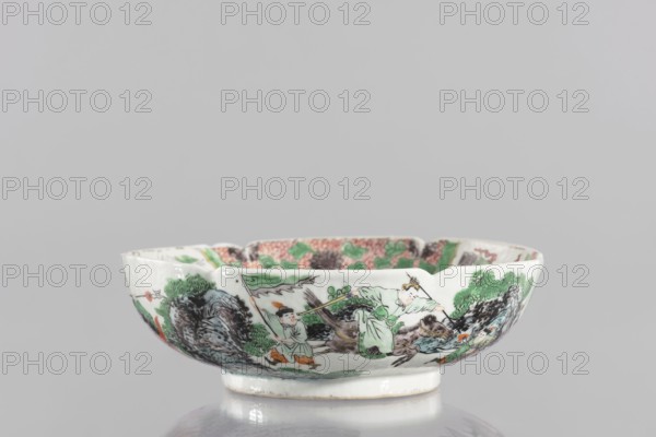 Bowl, ceramics, figural scenes, knightly scenes, Chinese art, colored porcelain, backdated signatures, six-leafed, bottled belly. Porcelain body; white, transparent glaze. Decoration painted with overglaze enamel paints and iron red. 19th century, China. Vessel; height 6.5 cm, diameter 20 cm., A shallow porcelain bowl with a scalloped rim sits on a reflective surface; the bowl is primarily white with painted scenes around its exterior featuring figures and landscape elements in green, black, brown, orange, and a few touches of pink, with green foliage and dark rock-like shapes, and additional green and pink patterned decoration visible along the interior rim.
