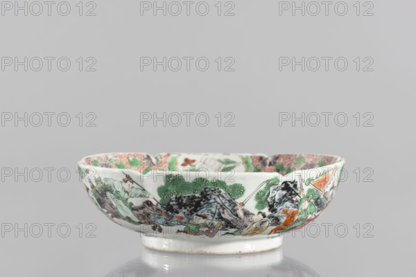 Bowl, ceramics, figural scenes, knightly scenes, Chinese art, colored porcelain with backdated signatures, six-leafed, bottled belly. Porcelain body; white, transparent glaze. Decoration painted with overglaze enamel paints and iron red. 19th century, China. Vessel; height 6.5 cm, diameter 20 cm., A shallow ceramic bowl with a scalloped rim sits against a neutral gray background, decorated with a painted scene featuring a group of figures amid stylized rocks and foliage; the decoration uses green, orange, black, brown, and pale blue pigments on a white ground, with several figures wearing green and orange garments and black hair details, leafy green trees and dark mottled rocks forming a central landscape motif, and the interior rim showing additional colored patterns visible from the vantage point.