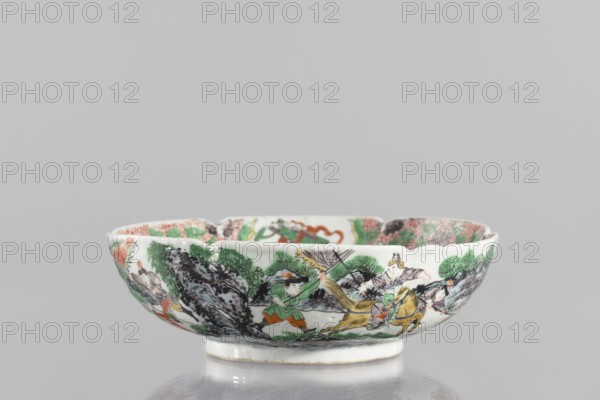 Bowl, ceramics; porcelain body with white, transparent glaze; decoration painted with overglaze enamel paints and iron red depicting figural and knightly scenes; colored porcelain with backdated signatures; six-leafed, bottled belly form. 19th century, China; vessel. Height 6.5 cm; diameter 20 cm., A shallow scalloped porcelain bowl with a glossy white base decorated around its exterior with a painted scene in green, orange, yellow, black, gray, brown, and pink tones showing a group of figures amid stylized rocks and foliage; one figure appears to wear green clothing, another is seated on a yellow animal, and several others are arranged along the band; the interior rim shows repeating decorative elements in similar colors, and the bowl sits on a short circular foot against a light gray background.