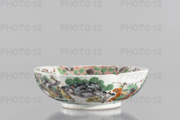 Bowl, Porcelain body; white, transparent glaze. Decoration painted with overglaze enamel paints and iron red, colored porcelain, figural scenes, knightly scenes. Backdated signatures. Six-leafed, bottled belly. 19th century, China. Vessel; height 6.5 cm, diameter 20 cm., A shallow ceramic bowl sits on a reflective surface, showing a curved white exterior decorated with a painted scene of people and animals amid stylized trees and rocks; the decoration includes greens, oranges, yellows, blacks, browns, and subtle touches of red, with figures wearing robes and headpieces and some animals depicted in motion, while the interior rim reveals additional patterned motifs in green, pink, and black.