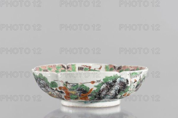 Bowl, gift, ceramics, figural scenes, knightly scenes, Chinese art, colored porcelain, backdated signatures, six-leafed, bottled belly. Porcelain body; white, transparent glaze. Decoration painted with overglaze enamel paints and iron red. 19th century, China, vessel. Height 6.5 cm; diameter 20 cm., A shallow ceramic bowl with a scalloped rim decorated in painted scenes showing a group of figures and landscape elements in colors including green, orange, black, brown, and blue, with foliage and rock-like patterns around the exterior and a glazed white interior and foot.