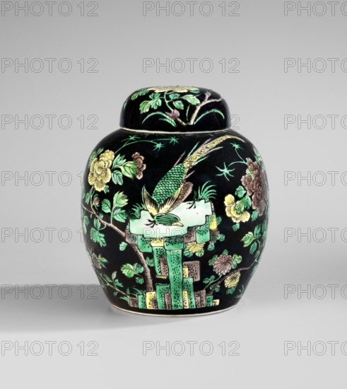 Ginger vessel with lid, porcelain pot decorated with overglaze enamel paints on a black background; Chinese, Qing dynasty 1644–1911, 19th century. Decoration includes pheasants, images of birds, rocks; colored porcelain spice vessel for ginger; height 15 cm, diameter 13.2 cm., A rounded ceramic jar with a lid sits against a light gray background casting a soft shadow; the jar has a glossy black ground decorated with floral and bird motifs in green, yellow, brown, and pale turquoise; the lid mirrors the black background and shows green leaves and flower elements; the body features a stylized bird with green and yellow plumage perched on a pale turquoise rock-like form, surrounded by branches with green leaves and yellow and brown blossoms, and there are geometric vertical elements in shades of green and pale yellow near the base.