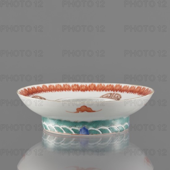 Bowl stand. Porcelain shard in the form of a ring with white glaze. Decoration painted with overglaze iron red, blue and green enamel, including bats and shou longevity hieroglyphs. Colored porcelain stand, Qing dynasty, 19th century, China. Height 3.3 cm; diameter 12.5 cm., Classification: Chinese ceramics., A shallow ceramic dish with a flared rim resting on a short foot, shown against a gray background. The upper surface of the dish is glossy white with a decorative band of reddish-orange petal-like shapes around the inner rim and a small reddish-orange cloud motif on the outer wall. The foot is painted in shades of green with a repeating wave pattern outlined in white, and there is a small blue accent where the foot meets the base. The edge of the rim has a thin warm-toned line, and the dish sits on a reflective surface that mirrors its colors. The visible colors include white, reddish-orange, green, blue, warm brown, and gray.