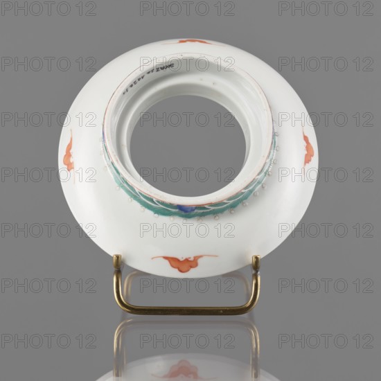 Bowl stand. Porcelain shard in the form of a ring with white glaze; decoration painted with overglaze iron red, blue and green enamel showing bats and shou longevity character/hieroglyphs. Colored porcelain, Qing dynasty, 19th century, China. Height 3.3 cm; diameter 12.5 cm., A round white ceramic object with a central circular opening sits on a brass stand; the rim around the opening has a decorative band in green and blue with small raised white bead-like dots, and there are a few orange-red cloud-shaped motifs spaced on the white surface; the brass stand and the object cast a reflection on a gray surface and the background is gray.