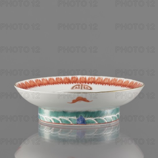 Bowl stand., Porcelain body with white glaze; decoration painted with overglaze iron red, blue and green enamel and gilding. In the form of a ring. Features iconography of bats and longevity shou character. Colored porcelain, Chinese; 19th century Qing dynasty, China. Vessel. Height 2.5 cm; diameter 12.5 cm., A shallow white porcelain dish with a flared rim sits on a short cylindrical foot; the rim interior is decorated with a repeating orange-red petal-like pattern and the dish interior shows a small orange-red decorative motif near the center; the exterior rim edge has a thin metallic-looking band and a small orange-red accent on the side; the foot is painted with a band of green brushwork and a repeating scalloped white-and-green wave pattern punctuated by a small blue triangular element, and the whole object rests on a reflective gray surface under even lighting showing subtle reflections; the visible colors include white, orange-red, green, blue, gray, and a metallic gold-like ton...