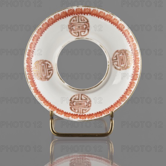Bowl stand, ; porcelain ring-form stand with white glaze; decoration painted with overglaze iron red, blue and green enamel and gilding; motifs: bowls, bats, longevity shou character; colored porcelain; hieroglyphs; 19th century Qing dynasty, China; vessel; height 2.5 cm, diameter 12.5 cm., A circular white ceramic ring with a central open hole sits upright on a small metallic stand; the rim features a repeating decorative border in a reddish-orange hue and several round medallion-like motifs in the same reddish-orange with gold outlining, while the stand is metallic gold and the background is a flat gray, and a faint reflection of the object appears below it.