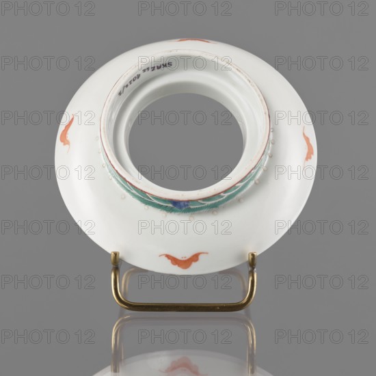 Bowl stand. In the form of a ring. Porcelain body; white glaze. Decoration painted with overglaze iron red, blue and green enamel, gilding; motifs include bats and longevity shou hieroglyphs. Colored porcelain, Chinese; 19th century Qing dynasty, China. Vessel. Height 2.5 cm; diameter 12.5 cm., A round white ceramic object with a central circular opening sits on a brass-colored U-shaped stand, showing a decorative band around the inner rim in teal and blue with small raised white dots; the white surface bears a few orange-red abstract motifs spaced around its edge, and the background is a neutral gray with a subtle reflection of the object and stand.