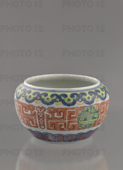 Globular vessel, porcelain body with white glaze. Decoration painted underglaze with cobalt, overglaze with enamel paints and iron red. Colored porcelain, longevity shou iconography, meander ornament. Low, flattened, without cover. Qing dynasty, 19th century, Chinese. Height 7.7 cm; diameter 10.3 cm; weight 0.35 kg., A small rounded ceramic vessel sits against a muted gray background, showing a pale interior and a glossy white exterior decorated with colorful painted bands; the top band features a flowing scroll-like pattern in shades of blue, yellow, and green, a central band displays geometric maze-like motifs and circular medallion shapes in orange and green, and the lower band has an arched pattern in blue and orange, with the overall colors visible including white, blue, yellow, green, and orange.