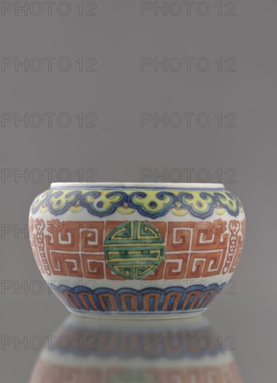 Globular vessel, porcelain body with white glaze; decoration painted underglaze with cobalt and overglaze with enamel paints and iron red. Low, flattened form without cover. Colored porcelain, Chinese, Qing dynasty 19th century. Decoration includes longevity shou iconography and meander ornament. Height 7.7 cm; diameter 10.3 cm; weight 0.35 kg., A rounded ceramic bowl with a glossy finish sits against a neutral gray background, showing a palette of white, blue, green, yellow, orange, and red; the rim area features a wavelike decorative band in blue and yellow, below which a central band displays repeating geometric motifs in orange interspersed with circular medallions in green; the lower section has an arched scallop pattern in blue and orange above a pale base, and a subtle reflection of the bowl appears on the surface beneath it.
