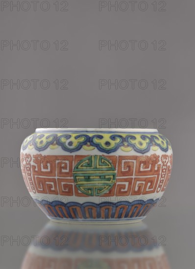Globular vessel, porcelain body with white glaze; decoration painted underglaze with cobalt, overglaze with enamel paints and iron red. Low, flattened, without cover. Colored porcelain with longevity shou iconography and meander ornament. Chinese, Qing dynasty 1644–1911, 19th century. Height 7.7 cm; diameter 10.3 cm; weight 0.35 kg., A small round ceramic bowl sits centered against a plain gray background, showing a smooth white base with decorative painted bands; the upper rim band features a wavy motif in yellow, blue, and green, while the central band displays a repeating geometric pattern in orange with an inset circular medallion in green and turquoise; the lower band shows an arch-like pattern in blue and orange above a thin pale base, and the bowl's glossy surface creates a soft mirrored reflection beneath it.