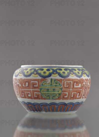 Globular vessel, porcelain body with white glaze; decoration painted underglaze with cobalt, overglaze with enamel paints and iron red. Low, flattened, without cover. Colored porcelain, Chinese, Qing dynasty 19th century. Motifs include longevity shou iconography and meander ornament. Height 7.7 cm; diameter 10.3 cm; weight 0.35 kg., A small rounded ceramic bowl sits against a plain gray background, showing a glossy white base with decorative bands in blue, green, yellow, red, and orange; the upper rim area features a scalloped cloud-like pattern in blue, yellow, and green, a central band displays repeating geometric motifs in red-orange with a circular medallion in green, and the lower edge shows alternating arch shapes in blue and red-orange, with the bowl and its colorful patterns reflected softly on the surface below.