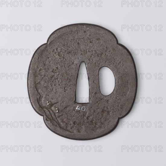 Crossguard tsuba, Kaneie,  school craftsman author. Japanese, Edo period 1603–1868, 18th century. Tsuba in the shape of a quatrefoil mokkô with an uchikaeshi edge, katahitsu ana one side hole. Decorated in relief showing the sennin Gama sitting under a tree, holding a staff and looking at a three-legged frog; on the reverse a sketched landscape motif with a waterfall. Details eyes, pebbles, staff inlaid with gold and shakudo. Origin: Fushimi, Yamashiro Province. Materials/subject: weapons — sword crossguard katana. Dimensions: height 7.2 cm, width 6.8 cm, depth 0.4 cm., A round, slightly irregular dark brown metal object with a textured, pitted surface and a scalloped outline, featuring two openings near the center—one elongated teardrop-shaped and one oval—and small raised decorative elements on one side with tiny pale inlays; the background is light gray and the visible colors include dark brown, pale off-white, and light gray.