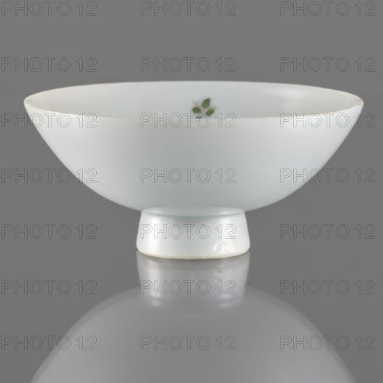 Bowl on a high foot, Porcelain bowl with white glaze and overglaze painted decoration of birds and flowers in huaniao style. Colored porcelain, Qing dynasty 1644–1911, 19th century. China. Vessel, height 4.2 cm, diameter 10.6 cm., A small shallow bowl with a rounded shape and a short cylindrical foot sits on a reflective surface, showing a soft reflection beneath it; the bowl is smooth and primarily white with a subtle glossy finish, and there is a small cluster of green leaf shapes visible on the inner wall; the background and reflection appear gray.