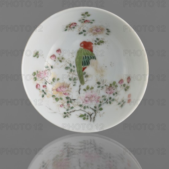 Bowl on a high foot, Chinese art, Chinese culture, Qing 1644–1911, Qing style, ceramics, colored porcelain, bowl with birds and flowers in huaniao style, porcelain, white glaze, overglaze painted decoration, 19th century Qing dynasty, China, vessel, height 4.2 cm, diameter 10.6 cm, A white porcelain dish viewed from above shows a painted scene of a perched bird and flowering branches; the bird has a red head and green and darker green wings, and it sits among branches with green leaves and several pink blossoms with touches of yellow and gold speckles; there are a few buds and small leaves scattered around the scene; the dish rests on a gray background and its painted image is faintly reflected below, showing muted pale pinks, greens, and grays.