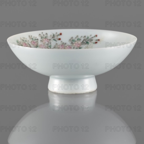 Bowl on a high foot, ceramics, bowls, Qing style, images of birds, Chinese art, colored porcelain, Chinese culture, huaniao style, Qing 1644–1911, porcelain pottery, white glaze. Decoration painted with enamel paints and overglaze iron red. 19th century, China, vessel; height 5.4 cm, diameter 11 cm., A shallow white ceramic bowl with a short cylindrical foot sits on a reflective gray surface, showing a soft mirror image beneath; the bowl's exterior is smooth white while the interior rim displays a floral pattern with pale pink blossoms, green leaves, and small red accents, all set against the white background, under even lighting and a neutral gray backdrop.