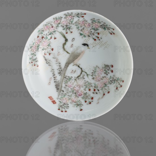 Bowl on a high foot, ceramics; bowl with painted images of birds; colored porcelain with white glaze; decoration painted with enamel paints and overglaze iron red; huaniao style, Qing 1644–1911, 19th century, China; porcelain vessel; height 5.4 cm, diameter 11 cm., A round white porcelain dish with a painted scene of a slender bird perched on a branch surrounded by flowering branches, a group of pale pink blossoms with green leaves and small red buds scattered around the composition; the bird has a black head, pale gray body, and muted brownish tail, and the branches are rendered in shades of green and brown with touches of gold speckling; the dish sits against a gray background and its lower edge and painted design are reflected faintly below, showing similar pale pink, green, red, white, gray, brown, black, and gold colors.
