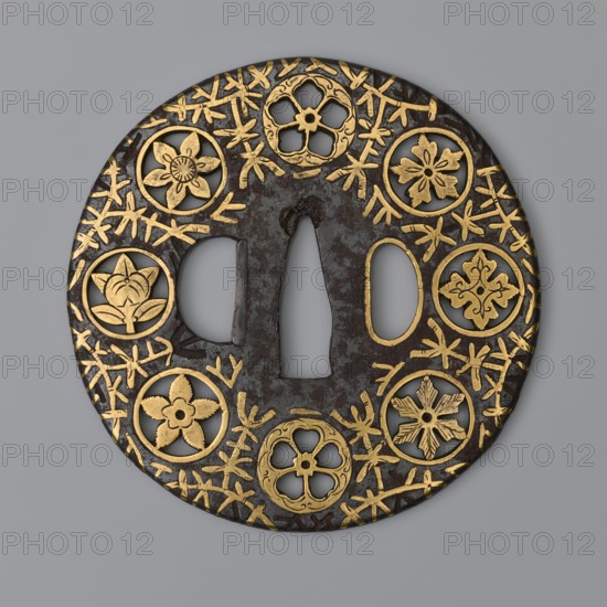 Round tsuba maru gata of the mon-sukashi type, decorated with eight openwork coats of arms monami arranged around the edge against a background of a brass-inlaid stylized vine karakusa. Crossguard tsuba, Japanese, katana accessory, edged weapon. 17th century Edo period, 1603–1868. Material: metal with brass inlay. Diameter 8 cm; thickness 0.3 cm., A round metal object with a dark brown surface decorated with a pattern of gold inlaid floral medallions and interlacing twig-like lines, featuring a central vertical opening flanked by two smaller openings, and several circular cutouts each containing different stylized flower or petal motifs in gold on the dark background; visible colors include dark brown, gold, and a neutral gray background.